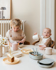 Two toddlers sit at a table with cake, drinks, and pastel plates. One eats while the other smiles at the camera, holding a mushie Snack Cup. A vase of daisies adds a cheerful touch to the scene.