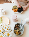 A child’s hand reaches for a Mushie Trainer Sippy Cup next to fruit, nuts, and daisies on a softly lit table, creating a bright, healthy snack time atmosphere.