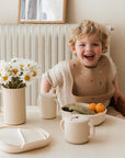 A laughing toddler sits at the table with a beige rocket-print bib, mushie cream children’s dinnerware, a spill-proof mushie Snack Cup, fresh fruit, and white daisies—perfect for happy little ones.