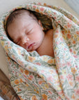 A newborn peacefully sleeps, wrapped in a mushie Organic Cotton Muslin Swaddle Blanket with soft yellow, pink, and green floral patterns, lying on a hospital bed.