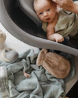 A baby in a gray tub is bathed as an adult supports their head, with a Mushie Organic Cotton Bath Mitt on a light blue towel nearby—ideal for washing delicate hair and sensitive skin.