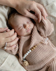 A baby sleeps peacefully on a mushie Organic Cotton Muslin Swaddle Blanket, wearing a pink knitted outfit with a beaded pacifier clip attached, while an adult gently holds the baby's hand and touches their head.