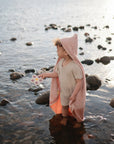 A young child stands in shallow water near the shore, wearing a beige outfit and a pink hooded towel. The child holds a mushie Flower Press Toy, exploring their senses as sunlight sparkles on the surface.