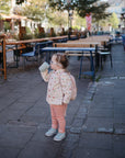 A young child in a mask, patterned pants, and a light jacket drinks from the Mushie Trainer Sippy Cup with easy-grip handles while standing on a paved area surrounded by empty tables, chairs, and trees in the background.