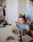 A toddler with pigtails sits at a table as an adult ties her mushie Silicone Baby Bib. Non-toxic silicone tableware and toys are scattered on the table in a bright, modern kitchen.