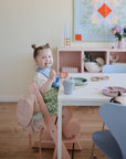 A young child wearing a green checkered dress and bib smiles while eating with a spoon at the table, sipping from the non-toxic Mushie Trainer Sippy Cup. A pink backpack, quilt, and flowers decorate the background.