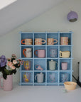 A pastel blue mug rack on a white counter holds colorful mugs and a Mushie Trainer Sippy Cup with easy-grip handles. A vase of flowers, coffee machine, and lilac wall lamp complete this organized kitchen corner.