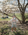 A toddler in a pastel outfit stands barefoot on grass, sipping from a cup under a blooming tree. Nearby, the mushie Silicone Pacifier Clip | Hera with its minimalist design is visible, while white flowers and blurred people suggest a springtime scene.
