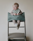 A smiling toddler in striped pajamas sits safely in a high chair, wearing a mushie Muslin Bib made of organic cotton; secured by a green fabric wrap against a plain, light background.