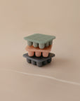 Three mushie Fresh Food Feeder Freezer Trays in muted green, pink, and gray are stacked against a beige background—perfect for convenient baby food storage.
