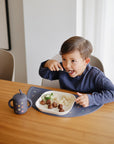 A child eats at a table next to a Mushie Silicone Training Cup + Straw.