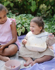 Two young children sit on a blanket outdoors. The older one smiles as the younger laughs, holding a mushie Silicone Baby Bib with an adjustable neck strap. Green foliage forms the backdrop.