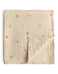 The mushie Organic Cotton Muslin Swaddle Blanket in Rainbows features a beige color, subtle stitched texture, and small brown rainbow patterns. One corner is folded to show the soft reverse side. Made from organic cotton for comfort.