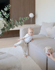 A toddler in light clothing sits on a beige sofa, reaching for a mushie Snack Cup held by an adult. The modern living room features neutral tones, a round table, and a vase with green foliage.