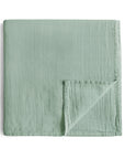 The mushie Organic Cotton Muslin Swaddle Blanket in Roman Green features a neatly folded design with a stitched grid pattern and a turned-over corner, displayed on a white background.