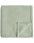 The mushie Organic Cotton Muslin Swaddle Blanket in Sage is shown neatly folded on a white background, with one corner flipped to reveal its grid pattern, organic cotton texture, and detailed stitching.