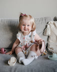 A smiling toddler with light hair and a pink clip sits on a gray couch, holding mushie Bath Boats. She wears a white dress with hearts and knee-high socks, surrounded by other colorful water play toys.