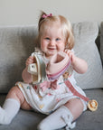 A smiling baby with light hair and a pink bow sits on a gray sofa, holding pastel stacking toys and the FRIGG Daisy Natural Rubber Pacifier by FRIGG. She wears a white dress with colorful shapes and white knee-high socks.