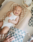 A smiling baby in a white onesie with a floral headband lies on Mushie Changing Pad Liners, surrounded by checkered accessories and baby items, looking happy and playful.