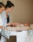A woman smiles at a baby on a changing table as an older child stands nearby, holding the mushie Water Resistant Wet Bag. The baby wears a striped hat in the softly lit, cozy room, highlighting this diaper bag storage moment.