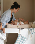A woman smiles and gently touches her baby's nose while changing them on the mushie Extra Soft Muslin Changing Pad Cover. A child watches nearby in a softly lit, cozy room with checkered cloth banners hanging from the table.