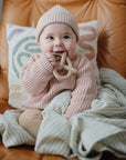 A baby in a pink knit outfit and hat sits on a tan leather chair, holding and chewing on the mushie Pretzel Teether, with a striped blanket and decorative pillow in the background.