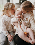 Three young children sit close together indoors. The older girl holds a baby using the mushie Silicone Pacifier Clip | Eva, while another girl kisses the baby's head. All wear light-colored clothing and share an affectionate moment.