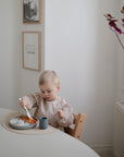 A young child with short blond hair eats rice and sauce with a spoon while wearing the mushie Long Sleeve Bib, which is water resistant. The minimalistic room has light walls, framed art, and a branch with purple leaves by the window.