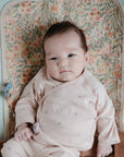 A baby in a pale pink outfit with a pacifier clipped on lies calmly on Mushie Changing Pad Liners with a floral pattern, looking at the camera. The background features soft pastel tones.