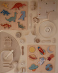 A flat lay of children's wooden dinosaur and space puzzles, silicone feeding utensils, a star teether, astronaut and rainbow toys, and the mushie Organic Cotton Muslin Swaddle Blanket on a cream-colored surface.
