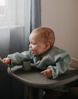 A smiling baby in a mushie Long Sleeve Bib sits in a high chair, playfully reaching out with a wooden spoon. A green cup is on the tray as sunlight streams through sheer curtains.