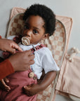 A toddler with a pacifier lies on a checkered changing pad as an adult adjusts their clothing. Mushie Changing Pad Liners and a folded pink cloth sit nearby with a small toy, while the child gazes up calmly.