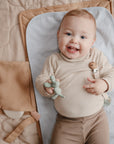 A smiling baby with light hair lies on a quilted surface, wearing a beige ribbed outfit and holding a green dinosaur toy. Nearby are a tan pouch, pacifier clip, and Mushie Changing Pad Liners.