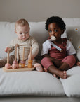 Two toddlers sit on a white couch. One plays with a wooden stacking toy, while the other, using a FRIGG Natural Rubber Pacifier 4-Pack by FRIGG, looks toward the camera. The couch has a pillow with white and yellow flowers.