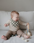 A smiling baby wearing a mushie Muslin Bib sits on a white couch, playing with a wooden stacking toy and scattered pieces.