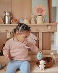 A young child with braided hair sits on a wooden bench in a sunlit room, wearing a pink sweater and blue pants. Next to her is the FRIGG Daisy Silicone Pacifier 2-Pack by FRIGG, placed among wooden toys and shelves.
