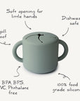 The mushie Snack Cup is a green silicone snack cup for toddlers with two handles and a soft slit lid, featuring spill-proof design, soft opening for little hands, dishwasher safe, and made from 100% food-grade silicone free from BPA/BPS/PVC/Phthalates.