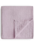 The mushie Organic Cotton Muslin Swaddle Blanket in Soft Mauve is neatly folded with one corner turned to reveal the reverse side, showing its subtle square pattern, all set against a white background.