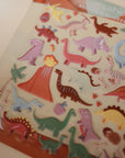 The mushie Reusable Sticker Set features colorful cartoon dinosaurs, a volcano, palm trees, and dino eggs on a beige background—perfect for fans of reusable sticker sets or sticker books.