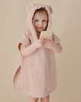 A young child smiles at the camera, holding the mushie Phone Press Toy—a beige sensory toy perfect for developing fine motor skills—while wrapped in a soft, light brown hooded towel with bear ears against a beige background.