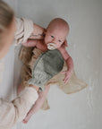 A caregiver bathes a baby in a tub, holding them with one arm while gently washing with a green cloth. The baby calms with the Mushie FRIGG Rope Natural Rubber Pacifier 2-Pack—one of many trusted baby essentials.