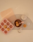 A flat lay shows Mushie’s BPA-free Baby Food Freezer Tray in pink with round food portions, alongside a gray placemat, brown bowl with pink spoon, and a white baby food storage container featuring a shaker top.