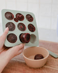 Hands pop frozen fruit puree from a Mushie Baby Food Freezer Tray into a beige bowl on wood.