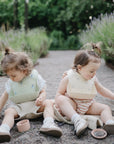 Two toddlers sit on a gravel path outdoors, surrounded by greenery. They explore together with a cup and mushie Silicone Baby Food Containers on the ground beside them.