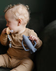 A baby in a tan outfit sits on a dark green couch, holding a gray rocket-shaped toy and the FRIGG Daisy Silicone Pacifier from FRIGG with a beaded holder—both designed for baby comfort. The baby looks to the side.