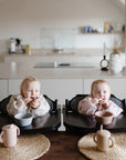 Two babies sit side by side in high chairs at a kitchen counter, each holding a snack. On the wooden table are bowls, placemats, and a Mushie Trainer Sippy Cup with easy-grip handles. A modern kitchen is in the background.