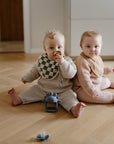Two babies sit back-to-back on a wooden floor, wearing knit outfits and mushie Muslin Bibs. One is chewing a teether toy, while a blue dinosaur rattle and pacifier are placed nearby.