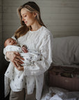 A woman in a white blouse gently cradles her newborn wrapped in a mushie Organic Cotton Muslin Swaddle Blanket, in a softly lit bedroom featuring white wooden walls and a pink diaper bag on the bed.