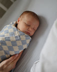 A newborn sleeps peacefully on a light-colored bed with the mushie Extra Soft Bassinet Muslin Crib Sheet, swaddled in a blue and white checkered blanket as an adult hand gently offers comfort.