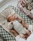 A baby in a cream-colored onesie lies on an organic cotton diaper changing pad while an adult gently holds them; Mushie Changing Pad Liners and folded baby clothes are organized nearby.